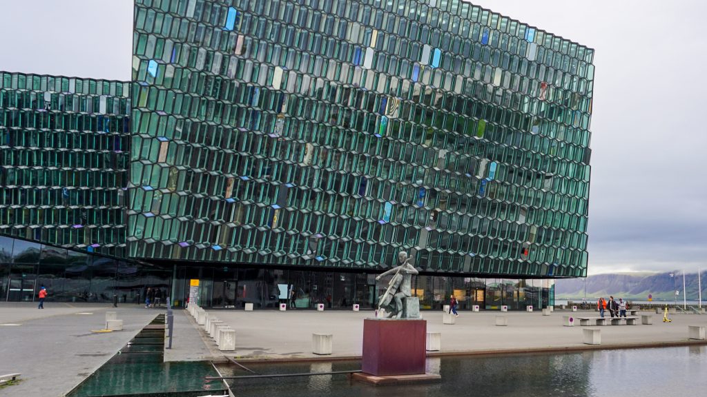 A glass building with green and blue tiles with a small fountain and a sculpture of violinist in front of it