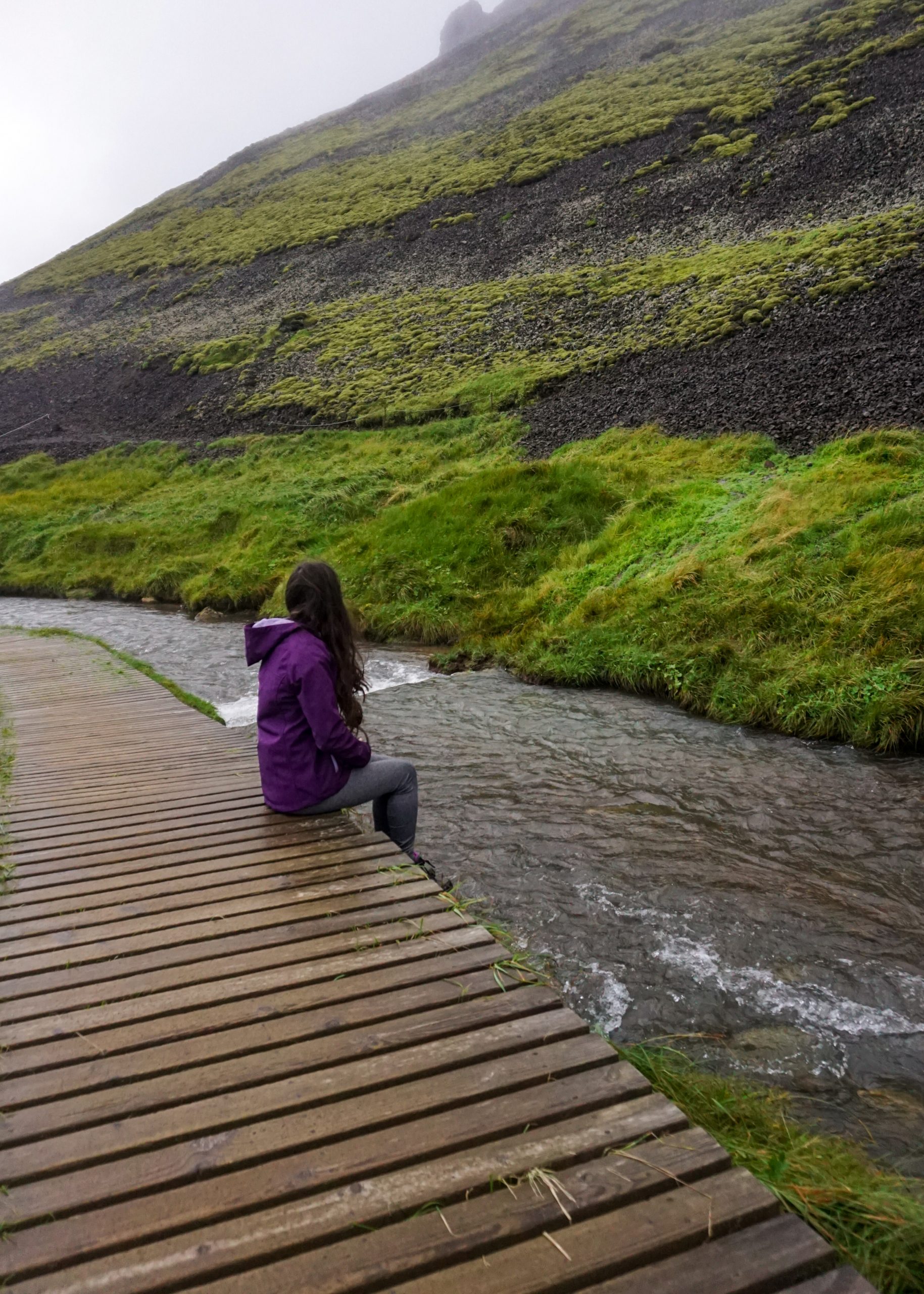 A girl in a purple jacket sitting near a hot spring in Reykjadalur Valley in Iceland