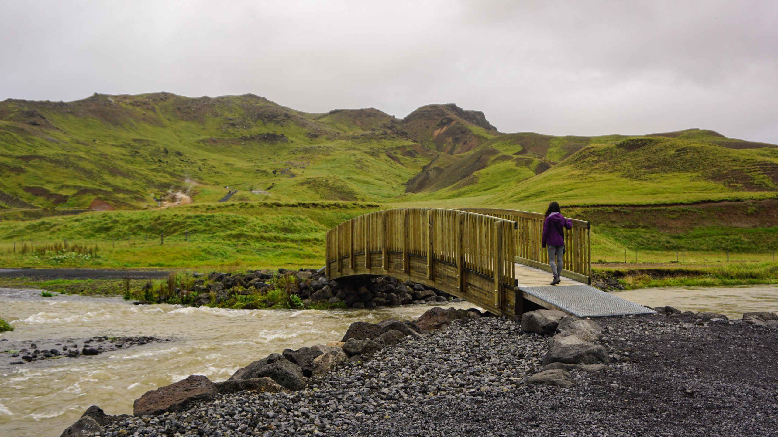 A girl in a purple jacket crossing a bridge in Reykjadalur Valley