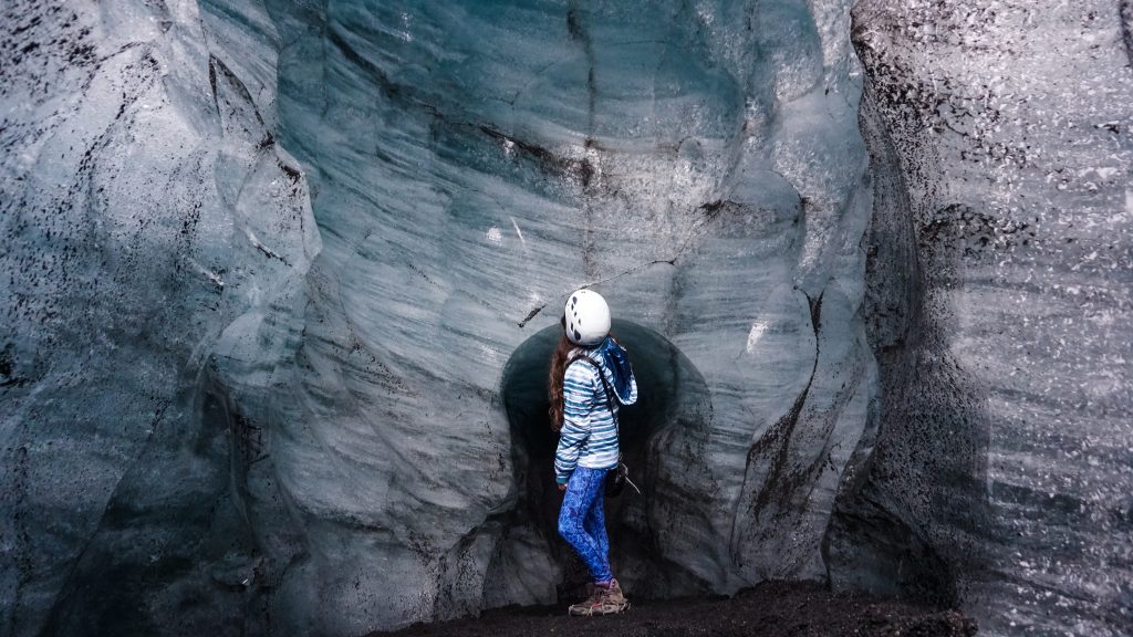 A girl wearing a blue outfit stands inside an ice cave wearing a white helmet and crampons on top of her shoes